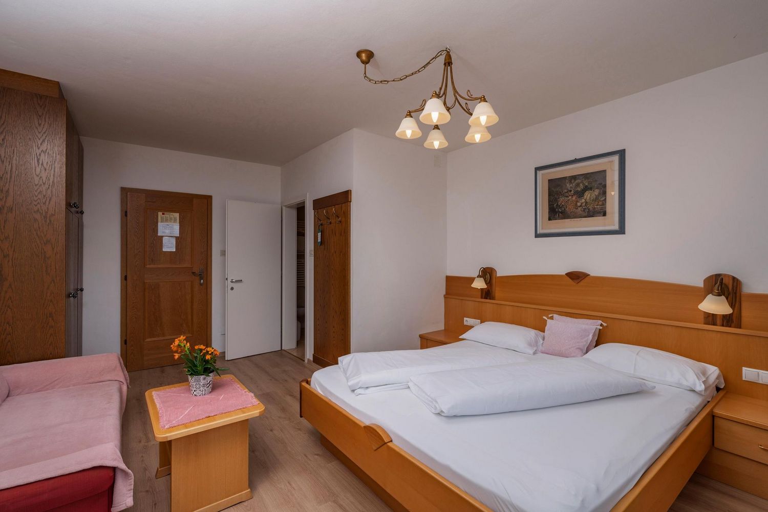 Ein gemütlich eingerichtetes Hotelzimmer mit einem Doppelbett aus hellem Holz, einem kleinen Tisch mit orangefarbenen Blumen, einem Sofa und einer klassischen Deckenleuchte.